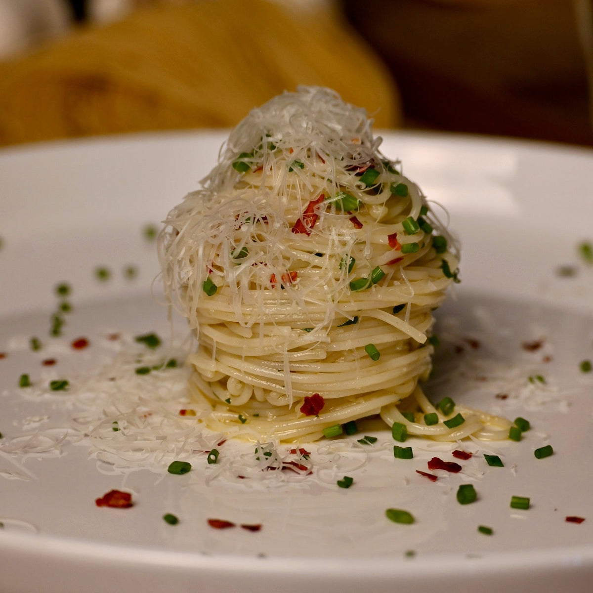A close-up shot of a gourmet dish of stacked Capellini Angel Hair noodles topped with shredded parmesan cheese, garnished with chopped chives and red pepper flakes, served on a white plate.