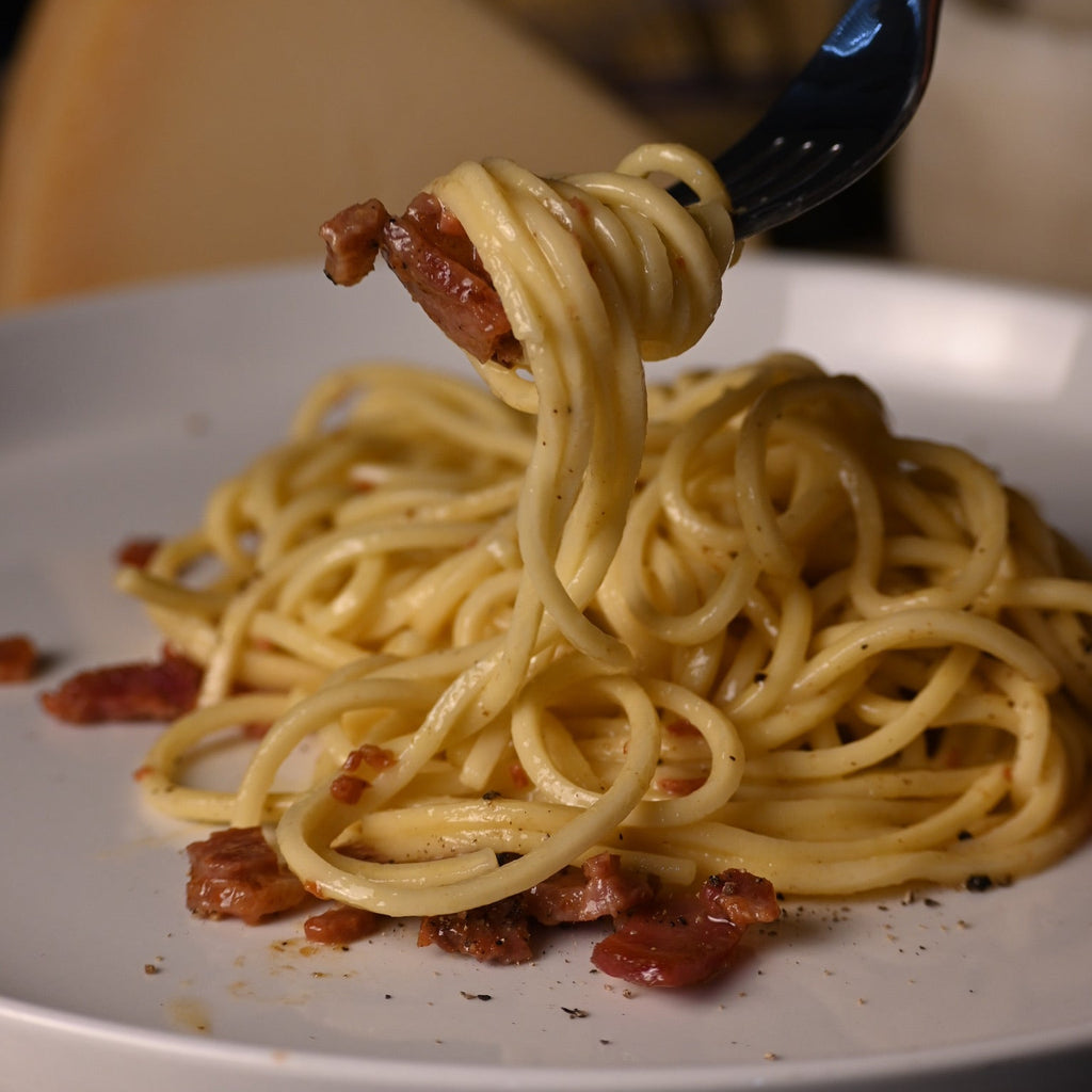 A close-up of a fork lifting a swirl of creamy linguine with pieces of cooked bacon from a white plate, with a wedge of cheese and dark background elements blurred behind it.