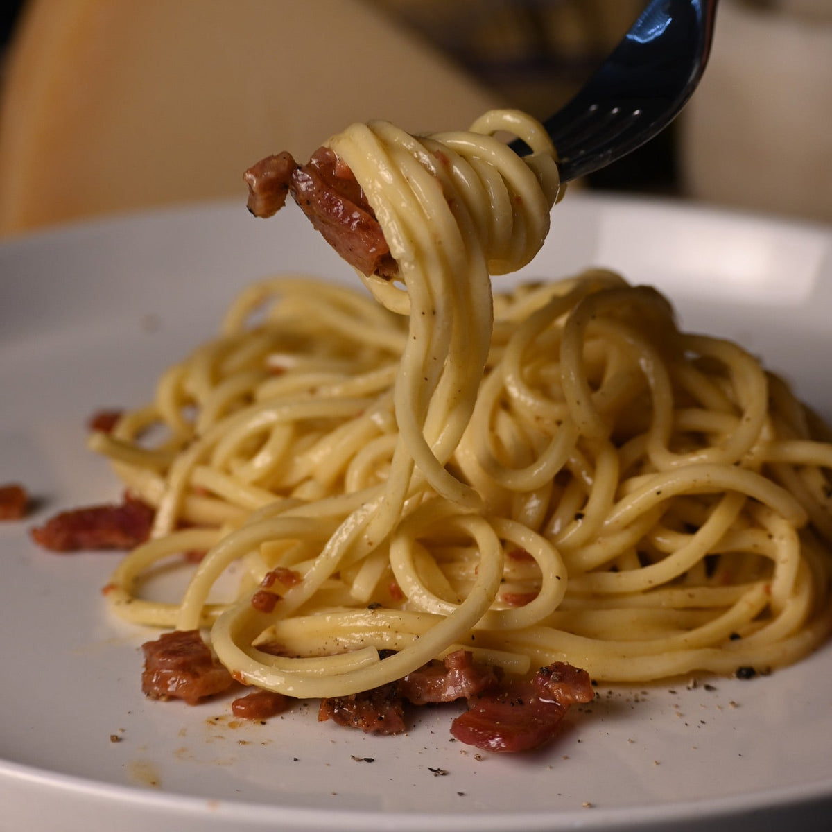 A close-up of a fork lifting a swirl of creamy linguine with pieces of cooked bacon from a white plate, with a wedge of cheese and dark background elements blurred behind it.