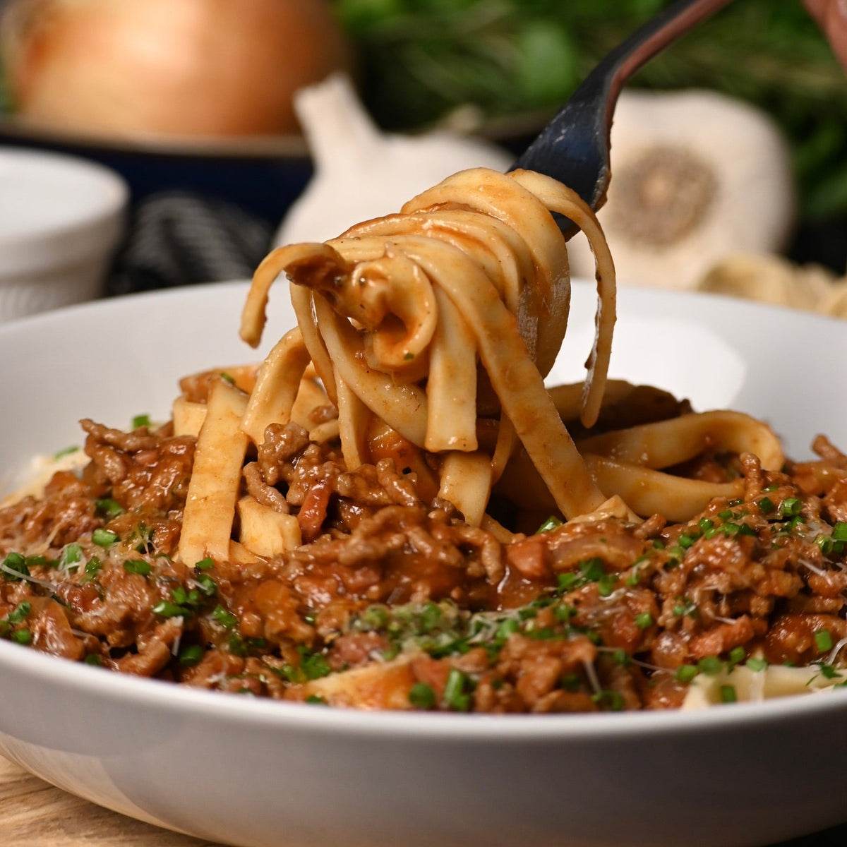 A close-up of a white bowl filled with pappardelle pasta and a meat sauce, with a hand using a fork to lift some of the pasta. A blurred onion, garlic, and green herbs sit in the background on a dark surface.