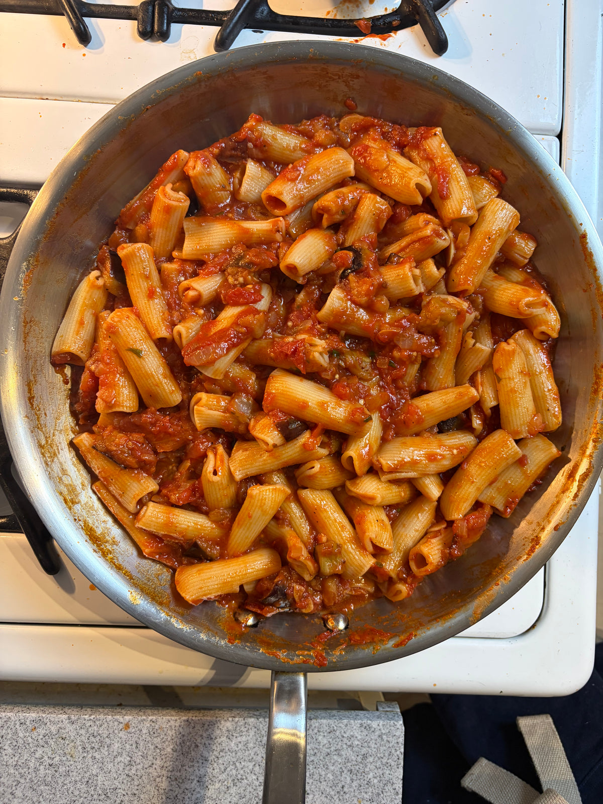 Rigatoni pasta in a stainless steel skillet with red tomato sauce, shown cooking on a white gas stovetop.
